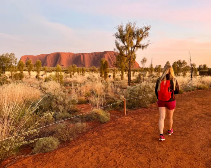 Kvinne går på en sti med Uluru-klippen i bakgrunnen mens solen står opp, Uluru-Kata Tjuta nasjonalpark, Australia